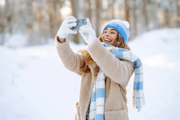 A young woman in a snowy park with a phone in her hands communicates via video and takes a selfie. Beautiful female tourist with phone having fun outdoors. Travel concept.