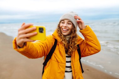 Sarı ceketli genç bir turistin portresi. Deniz kıyısında elinde telefon var. Güzel bir kadın telefonuyla selfie çekiyor, deniz havasının tadını çıkarıyor. Seyahat kavramı, blog yazma.