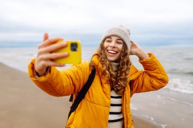Sarı ceketli genç bir turistin portresi. Deniz kıyısında elinde telefon var. Güzel bir kadın telefonuyla selfie çekiyor, deniz havasının tadını çıkarıyor. Seyahat kavramı, blog yazma.