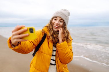 Sarı ceketli genç bir turistin portresi. Deniz kıyısında elinde telefon var. Güzel bir kadın telefonuyla selfie çekiyor, deniz havasının tadını çıkarıyor. Seyahat kavramı, blog yazma.