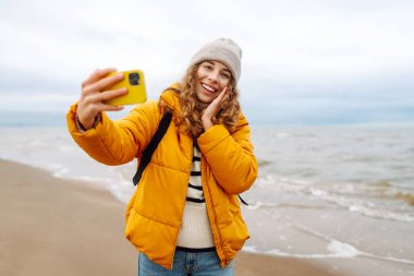Sarı ceketli genç bir turistin portresi. Deniz kıyısında elinde telefon var. Güzel bir kadın telefonuyla selfie çekiyor, deniz havasının tadını çıkarıyor. Seyahat kavramı, blog yazma.