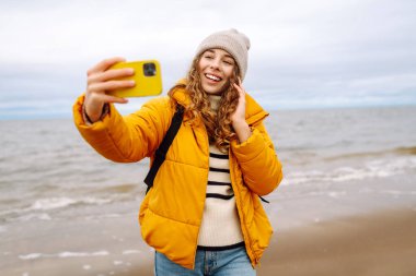 Sarı ceketli genç bir turistin portresi. Deniz kıyısında elinde telefon var. Güzel bir kadın telefonuyla selfie çekiyor, deniz havasının tadını çıkarıyor. Seyahat kavramı, blog yazma.