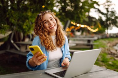 Telefonlu ve dizüstü bilgisayarlı genç kıvırcık kadın gölün arka planında oturuyor. Dişi serbest çalışan biri dışarıda bir dizüstü bilgisayarda çalışıyor. Teknoloji konsepti. Yaşam biçimi.