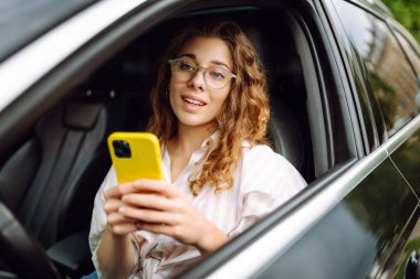 Elinde telefonla araba süren güzel kıvırcık bir kadının portresi. Mutlu kadın araba hareketleri için mobil navigasyon kullanıyor. Taşımacılık kavramı, hareketlilik.