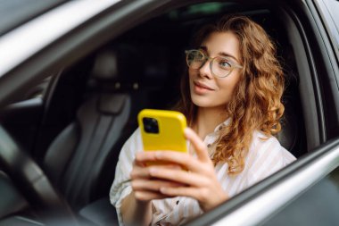 Elinde telefonla araba süren güzel kıvırcık bir kadının portresi. Mutlu kadın araba hareketleri için mobil navigasyon kullanıyor. Taşımacılık kavramı, hareketlilik.