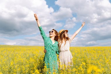 Neşeli kız arkadaşlar güneşli bir havada çiçek açan bir kolza tarlasında yürüyorlar. Dışarıda eğlenen ve eğlenen iki genç kadın. Doğa ve eğlence kavramı.