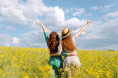 Neşeli kız arkadaşlar güneşli bir havada çiçek açan bir kolza tarlasında yürüyorlar. Dışarıda eğlenen ve eğlenen iki genç kadın. Doğa ve eğlence kavramı.