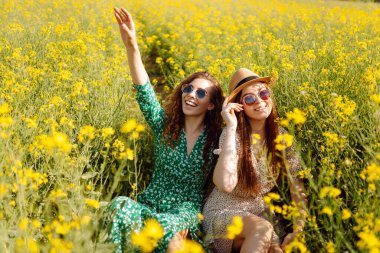 Neşeli kız arkadaşlar güneşli bir havada çiçek açan bir kolza tarlasında yürüyorlar. Dışarıda eğlenen ve eğlenen iki genç kadın. Doğa ve eğlence kavramı.
