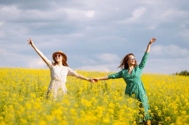 Neşeli kız arkadaşlar güneşli bir havada çiçek açan bir kolza tarlasında yürüyorlar. Dışarıda eğlenen ve eğlenen iki genç kadın. Doğa ve eğlence kavramı.
