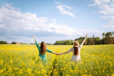 Neşeli kız arkadaşlar güneşli bir havada çiçek açan bir kolza tarlasında yürüyorlar. Dışarıda eğlenen ve eğlenen iki genç kadın. Doğa ve eğlence kavramı.