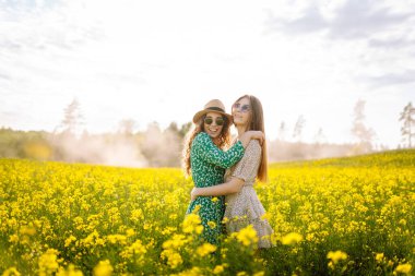 Neşeli kız arkadaşlar güneşli bir havada çiçek açan bir kolza tarlasında yürüyorlar. Dışarıda eğlenen ve eğlenen iki genç kadın. Doğa ve eğlence kavramı.