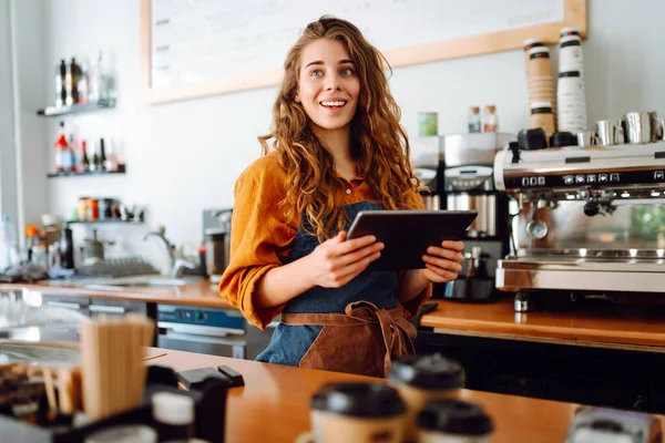 A young woman in an apron with a digital tablet at the bar counter of a coffee shop takes an order. Small business, technology concept.