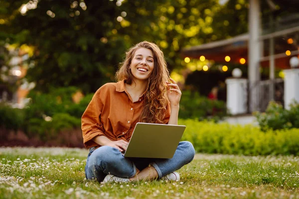 Happy woman with curly hair with a laptop on a green meadow at sunset. Young freelancer woman working outdoors. Freelance work, nature concept.