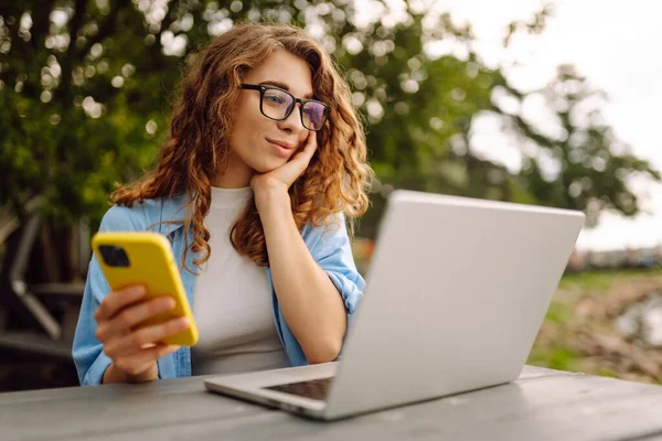 Young curly woman with a phone and laptop sitting at a table against the backdrop of a lake. Female freelancer works at a laptop outdoors. Technology concept. Lifestyle.