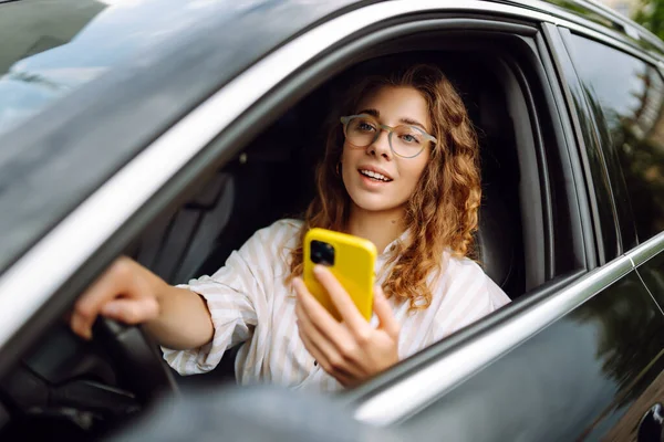 Portrait of a beautiful curly woman driving a car with a phone in her hands. Happy woman uses mobile navigation for car movement. Concept of transport, mobility.