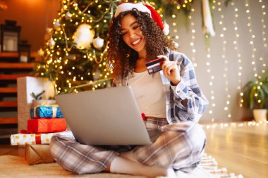 Haritası ve dizüstü bilgisayarı olan genç kıvırcık kadın Noel ağacının yanında oturuyor. Güzel bir kadın evde yeni yılın tadını çıkarıyor, hediyelerini paketliyor. İnternetten alışveriş, tatil konsepti.