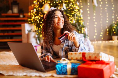 Haritası ve dizüstü bilgisayarı olan genç kıvırcık kadın Noel ağacının yanında oturuyor. Güzel bir kadın evde yeni yılın tadını çıkarıyor, hediyelerini paketliyor. İnternetten alışveriş, tatil konsepti.
