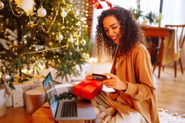 Haritası ve dizüstü bilgisayarı olan genç kıvırcık kadın Noel ağacının yanında oturuyor. Güzel bir kadın evde yeni yılın tadını çıkarıyor, hediyelerini paketliyor. İnternetten alışveriş, tatil konsepti.