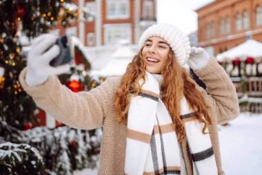 Güzel genç bir kadın yeni yıl fuarında eğleniyor ve selfie çekiyor. O bir kürk manto, şapka ve eşarp giydi. Harika bir yeni yıl havası. Tatil konsepti, hafta sonları, rahatlama.. 