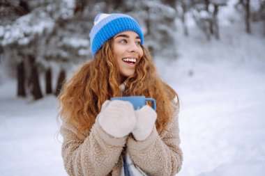 Elinde termos ve elinde bir fincan kahve olan genç bir kadın kış ormanındaki ilk karın tadını çıkarıyor. Tatil konsepti, doğa.