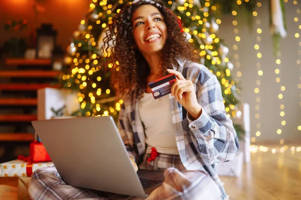 Young curly woman with a map and laptop sitting at home near the Christmas tree. Beautiful woman at home enjoying the New Year's atmosphere, packing gifts. Online shopping, holiday concept.