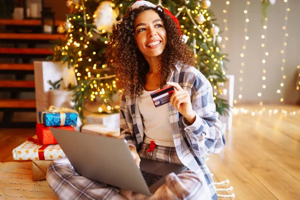 Young curly woman with a map and laptop sitting at home near the Christmas tree. Beautiful woman at home enjoying the New Year\'s atmosphere, packing gifts. Online shopping, holiday concept.