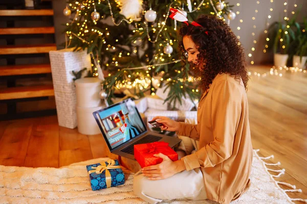 Young curly woman with a map and laptop sitting at home near the Christmas tree. Beautiful woman at home enjoying the New Year\'s atmosphere, packing gifts. Online shopping, holiday concept.