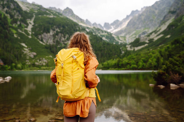 Beautiful female traveler with a yellow hiking backpack against the backdrop of a mountain lake with hiking poles. A young woman posing on a high mountain lake. Concept of freedom, tourism.