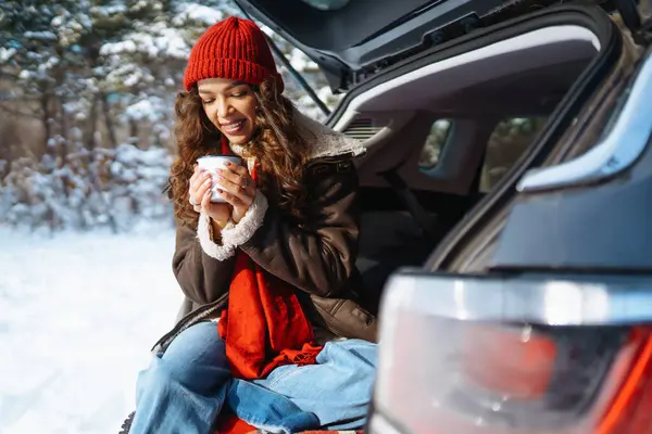 若い女性の旅行者は雪の道を見下ろす熱湯からホットドリンクを飲む車のトランクに座っています 美しい女性が屋外でリラックス 旅行コンセプト ライフスタイル — ストック写真
