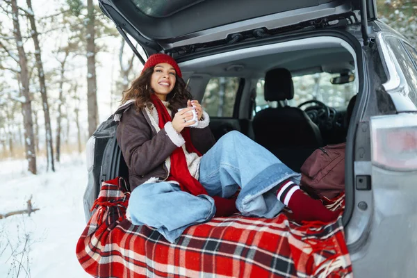 若い女性の旅行者は雪の道を見下ろす熱湯からホットドリンクを飲む車のトランクに座っています 美しい女性が屋外でリラックス 旅行コンセプト ライフスタイル — ストック写真