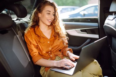 Güzel bir iş kadını bir arabanın arka koltuğunda otururken dizüstü bilgisayar kullanıyor. Arabada dizüstü bilgisayarla çalışan serbest çalışan genç bir kadın. Serbest, ulaşım kavramı.