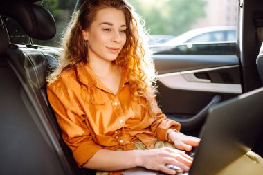 Güzel bir iş kadını bir arabanın arka koltuğunda otururken dizüstü bilgisayar kullanıyor. Arabada dizüstü bilgisayarla çalışan serbest çalışan genç bir kadın. Serbest, ulaşım kavramı.