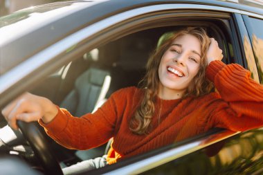Şehrin etrafında araba süren gülümseyen genç bir kadın. Kıvırcık saçlı kadın arabayla seyahat eder. Yaşam tarzı kavramı.