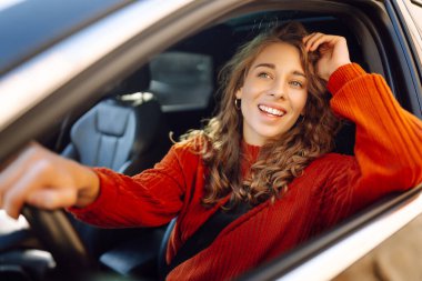 Şehrin etrafında araba süren gülümseyen genç bir kadın. Kıvırcık saçlı kadın arabayla seyahat eder. Yaşam tarzı kavramı.