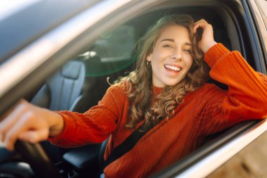 Şehrin etrafında araba süren gülümseyen genç bir kadın. Kıvırcık saçlı kadın arabayla seyahat eder. Yaşam tarzı kavramı.