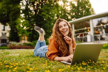 Kıvırcık saçlı, sıradan giyimli, dizüstü bilgisayarlı bir kadın güneşli bir günde çimlerin üzerinde oturuyor. Mutlu serbest çalışan kadın dışarıda çalışıyor ve havanın tadını çıkarıyor..