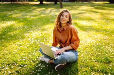 Kıvırcık saçlı, sıradan giyimli, dizüstü bilgisayarlı bir kadın güneşli bir günde çimlerin üzerinde oturuyor. Mutlu serbest çalışan kadın dışarıda çalışıyor ve havanın tadını çıkarıyor..