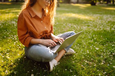 Kıvırcık saçlı, sıradan giyimli, dizüstü bilgisayarlı bir kadın güneşli bir günde çimlerin üzerinde oturuyor. Mutlu serbest çalışan kadın dışarıda çalışıyor ve havanın tadını çıkarıyor..