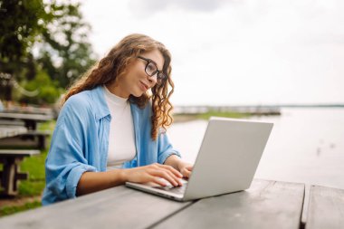 Göl kenarındaki açık bir kafede dizüstü bilgisayarlı genç bir kadının portresi. Kıvırcık kadın dışarıda dizüstü bilgisayarda çalışıyor. Serbest konsept, doğa.