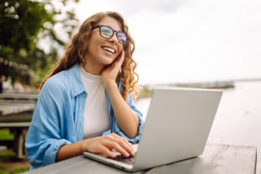 Göl kenarındaki açık bir kafede dizüstü bilgisayarlı genç bir kadının portresi. Kıvırcık kadın dışarıda dizüstü bilgisayarda çalışıyor. Serbest konsept, doğa.