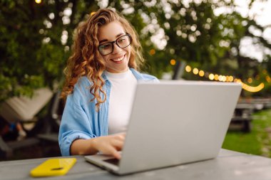 Göl kenarındaki açık bir kafede dizüstü bilgisayarlı genç bir kadının portresi. Kıvırcık kadın dışarıda dizüstü bilgisayarda çalışıyor. Serbest konsept, doğa.