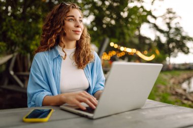 Göl kenarındaki açık bir kafede dizüstü bilgisayarlı genç bir kadının portresi. Kıvırcık kadın dışarıda dizüstü bilgisayarda çalışıyor. Serbest konsept, doğa.