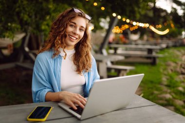 Göl kenarındaki açık bir kafede dizüstü bilgisayarlı genç bir kadının portresi. Kıvırcık kadın dışarıda dizüstü bilgisayarda çalışıyor. Serbest konsept, doğa.