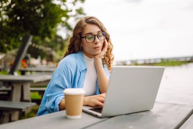 Göl kenarındaki açık bir kafede dizüstü bilgisayarlı genç bir kadının portresi. Kıvırcık kadın dışarıda dizüstü bilgisayarda çalışıyor. Serbest konsept, doğa.