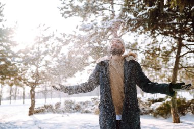 Genç sakallı bir adam karlı bir parkta yürüyor. Mutlu adam güneşli bir kış gününde havanın tadını çıkarıyor. Yürüyüş kavramı, hafta sonları.