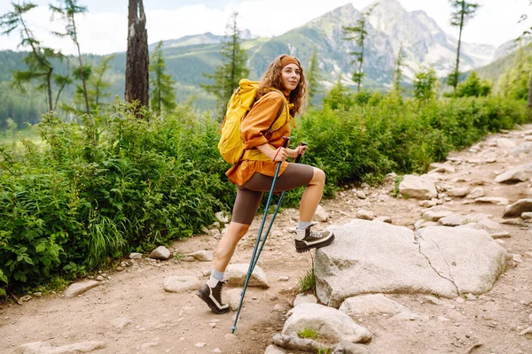 Young female traveler with hiking poles and a bright backpack enjoys the mountain scenery along a hiking trail. Active lifestyle. Wanderlust.
