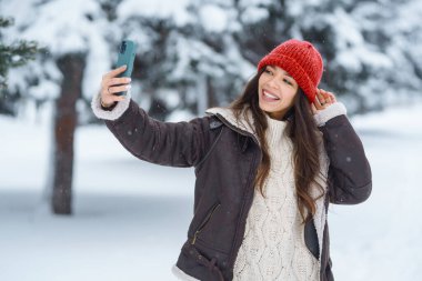 Parkta telefonuyla yürüyen, selfie çeken, blog yazan mutlu bir kadın. Genç bayan turist karlı bir parkta eğleniyor. Doğa kavramı, eğlence, rahatlama.