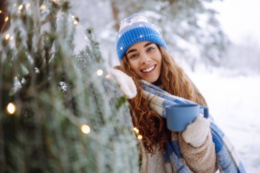 Genç bir kadın, karlı bir ormanda çelenkle süslenmiş bir Noel ağacıyla otururken termostan sıcak bir içecek içer. Gülümseyen kadın dışarıda Noel atmosferinin tadını çıkarıyor. Tatil kavramı.