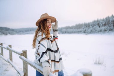 Kış ormanında kış gölü yakınlarında şapkalı ve atkılı mutlu bir kadın yürüyor. Kış yürüyüşü kavramı. Genç bir turist kadın açık havada soğuk bir günün tadını çıkarıyor..