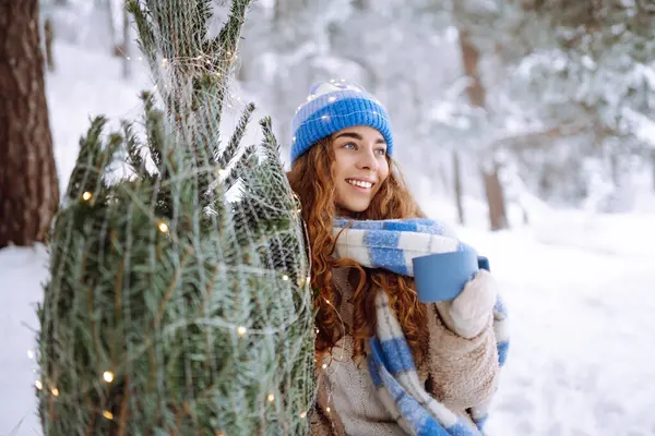 Genç bir kadın, karlı bir ormanda çelenkle süslenmiş bir Noel ağacıyla otururken termostan sıcak bir içecek içer. Gülümseyen kadın dışarıda Noel atmosferinin tadını çıkarıyor. Tatil kavramı.